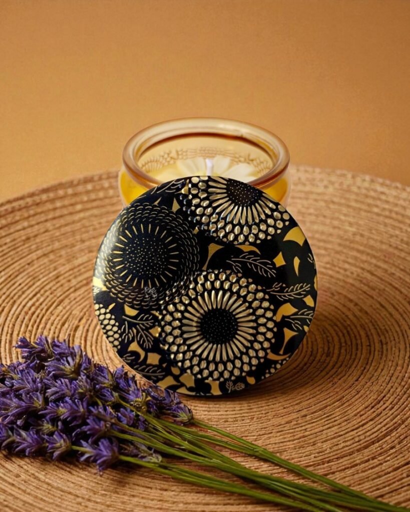 Vela aromática em pote de vidro âmbar com tampa decorada em preto e dourado com padrões florais, sobre uma base de palha trançada. Um pequeno ramo de lavanda roxa está posicionado ao lado da vela.