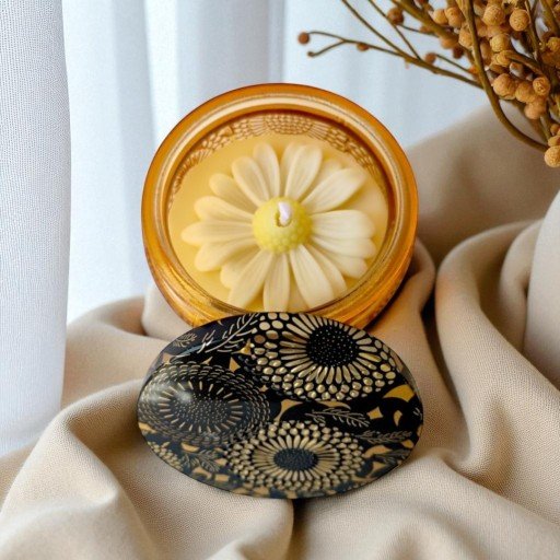 Vela decorativa em pote âmbar com flor de cera branca e tampa preta com desenhos florais dourados, apoiada sobre tecido bege, ao lado de ramos secos decorativos.