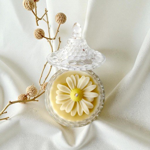 Vela artesanal em pote de cristal transparente com tampa trabalhada e cera amarela decorada com flor branca em relevo no topo, sobre tecido claro e ao lado de pequeno galho seco.